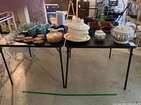 Two black card tables set side by side with various ceramic dishes and kitchenware placed on top, showing the full tops and legs.