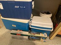 Three coolers stacked: blue Coleman Polylite 48 cooler on top, blue Thermos cooler below it, and white Rubbermaid cooler visible. The boxed dining canopy (12x12) is also present at the base of the stack with packaging stating "12x12 Dining Canopy" and showing canopy images.