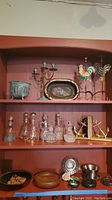 Multiple glass decanters, brass candlesticks, metal and ceramic decorative pots, trays, wooden bowls, metallic plate and bookends arranged on red shelves.