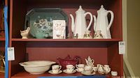 Overall view of shelf with coffee and tea pitchers, cookie jar, crock, serving bowl, cups and saucers, miniature tea set, and metal chairs arranged.