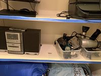 Shelf with RCA stereo system with two speakers, set beside plastic bins containing hair care appliances.