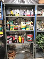 Four shelves with assorted garden and automotive liquid containers, small bottles, aerosol cans and flower pot trays on bottom shelf.