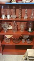 Full view of wooden shelves holding various glassware items including decanters, hurricane lights, pitchers, serving bowls, and hand fans.