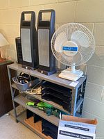 Image showing a shelving unit with two black tower space heaters, one white Holmes Air 3-speed oscillating fan, multiple black stackable paper trays, extension cords, umbrellas, and a gray cube organizer on bottom shelf.