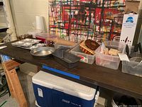 Photo showing metal bakeware including muffin tin, loaf pan, round cake pan, wooden rolling pin, plastic containers on a wooden shelf above a Coleman cooler