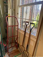 Hand dolly, two pitchforks and yard rake leaning against a window, next to ladders.