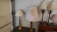 Four table lamps in a room against a garage door and next to furniture. One brass segmented column lamp with a cream shade, one wooden carved lamp with a pleated shade, and two black and gold accent lamps with cream shades visible.
