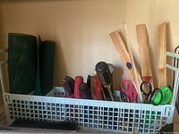 Photo shows a white plastic basket containing rolled green deer blocking mesh, hedge trimmers with green and black handles, wooden stakes, scissors, and assortment of hand tools.