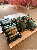 Photo of nine outdoor decorative pillows arranged on a white display surface showing a mix of floral striped pillows and solid green pillows with ruffled edges.