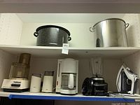 Shelf with large dark speckled enamel pot, stainless steel pot, white Cuisinart food processor, two coffee grinders, white Krups coffee maker, Hamilton Beach blender, and Rowenta iron.