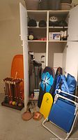 Photo showing croquet set with wooden mallets and colored balls on stand, orange sled, golf bag with clubs, boogie boards, camping chairs and helmets stored in white cabinet shelving.