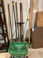 Full view of yard tools and Scotts fertilizer spreader leaning against wall indoors on a carpeted floor.