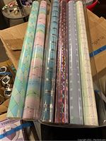 View of 5 rolls of wrapping paper showing pastel argyle, floral, plaid, iridescent, polka dots, and script designs.