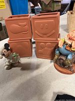 Four terracotta-colored ceramic square pots with different Mickey Mouse icons on each side and two Disney figurines: Christmas Mickey figurine holding a wreath and Snow White and the Seven Dwarfs Happy figurine sitting on a wood textured base.