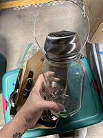 A clear glass mason jar with a metal handle and a silver-colored metal lid that includes a solar panel on top, held in hand.