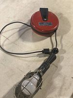 Full view of the hanging worklight with red retractable cord reel, black cord, and caged bulb light.