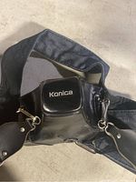 Black Konica Autoreflex camera in leather case with blue fabric camera strap attached.