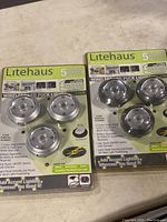 Two packages of Litehaus wireless LED puck lights, each package containing three lights in transparent and silver housing.