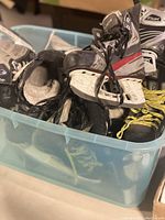 Side view of assortment of used hockey skates in a translucent plastic tote, showing varying colors and wear
