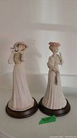 Two porcelain Victorian lady figurines on round wooden bases, showing entire set side-by-side.