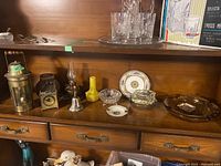 Display of assorted decorative items on a wooden shelf including oil lamps, desk clock, ashtrays, and porcelain items