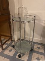 Full view of glass and metal bar cart with ice bucket on top, showcasing circular racks for six wine bottles on lower shelf.