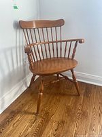 Single wooden turned wood chair with spindle backrest, armrests, and splayed legs on wood floor against white wall.