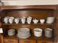 Full view of wooden cabinet shelf holding multiple teacups, saucers, bowls, and stacked plates including large dinner and smaller plates.