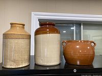 Three crocks displayed side by side on a black surface in front of a window, showing different heights and colors.