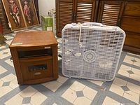 Photo showing wooden space heater and white box fan sitting side by side on tiled floor.