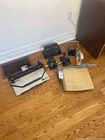 Wide shot showing all office supply items on hardwood floor including a JB International PB31SP wire binding machine, two black hole punches, white stapler, tape dispenser and a guillotine paper cutter.