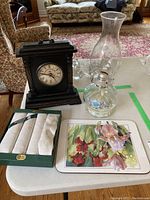 Photo showing all items on a table: black Colonial Clock Co. clock, clear glass oil lamp, boxed linen napkins, and iris floral hot plate mat.