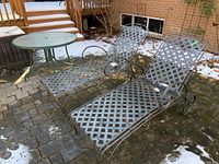 Two metal loungers with lattice pattern and wheels placed on stone patio, showing some rust and wear, view from side angle