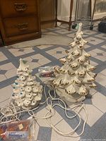 Two porcelain Christmas tree lamps side by side with cords and bags of colored bulbs on tiled floor