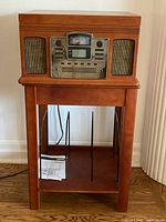 Front view of Crosley CR245 radio and CD recorder unit placed on wooden stand with manual on lower shelf, showing metal dividers and wood grain.