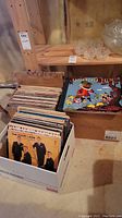 Three boxes of vinyl records stacked in a basement with wooden shelves and a cardboard sign labeled 'Records'. Visible album cover is The Four Lads.