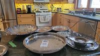 Overview of lot with multiple silver plated serving trays, a footed bowl, a handled basket, and a metal trivet arranged on kitchen counter.