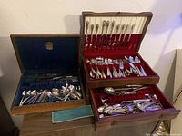 Two wooden boxes with silver plated cutlery displayed, showing various spoons, forks, knives and serving pieces inside with blue and red velvet lining