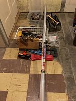 Photo of assorted tools and hardware in bins and tool bag on basement floor, with long metal power bar wrapped in packaging in front