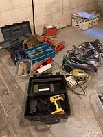 Overview of all tools and hardware including power drill in case, jigsaw, assorted hand tools, tool belts, extension cords, and power bars spread on basement floor.