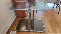 Metal framed trolley table with two tiers of smoky glass shelves shown against wooden floor and walls.