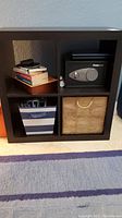 Full front view of black cubby shelving unit showing four compartments; two contain storage bins, one holds a safe and books.
