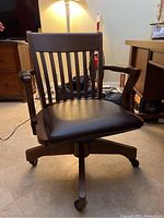 Front angle view of the wood swivel rolling desk chair showing vinyl padded seat, armrests, caster wheels and wooden backrest with vertical slats.