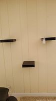 Three espresso wood floating shelves mounted on vertical paneling wall. Shelves are evenly spaced with two upper shelves and one lower shelf.