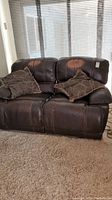 Full view of the espresso color leather reclining loveseat with two patterned pillows on a carpeted floor in front of a window.