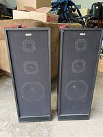 Front view of two RCA black floor-standing speakers with fabric grills, three speaker cones visible through the fabric on each.