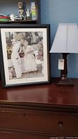 Framed wall art print and wood base table lamp positioned side by side on dark wood furniture against blue wall.