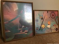 Two framed abstract art prints leaning against the wall, one slightly overlapping the other. Frames are faux wood with plexiglass protection. Prints have various geometric and flowing shapes.