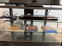 Wide view of shelf featuring Samsung Blu-ray player on top shelf with remotes and user manual, and multiple CDs and Blu-ray discs arranged below.