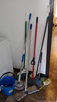 Photo showing multiple mops, brooms, and blue bucket against wall by laundry machine.